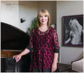Linda-In-Vegas-MagazineB A woman stands beside a grand piano, wearing a red and black patterned blouse, with a portrait in the background. The setting is elegant and calm, reflecting her role as a motivational speaker and disability advocate.| Linda’s Life – Linda Smith