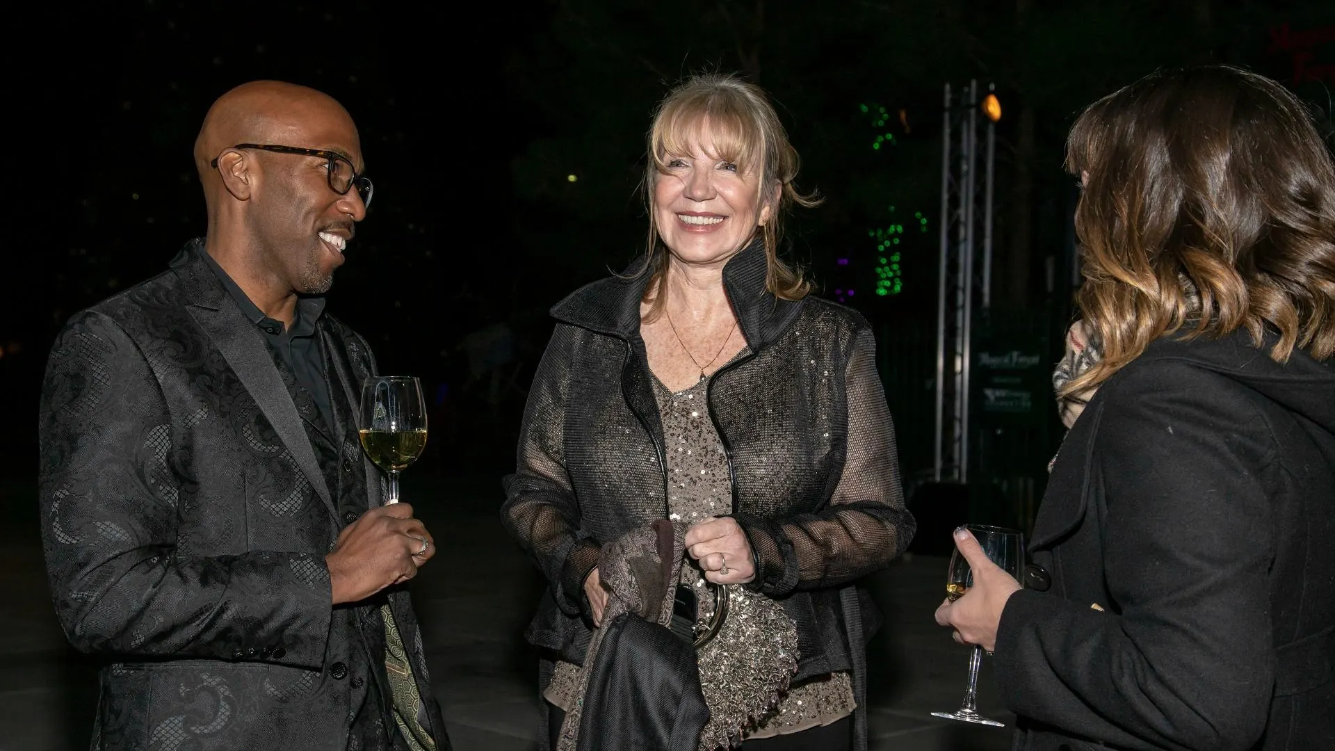 Linda Smith, an inspirational speaker, smiles while engaging with two others at a charity event. She wears a sparkly top and holds a jacket, embodying nonprofit leadership and advocacy in a warmly lit outdoor setting.| Linda’s Life – Linda Smith
