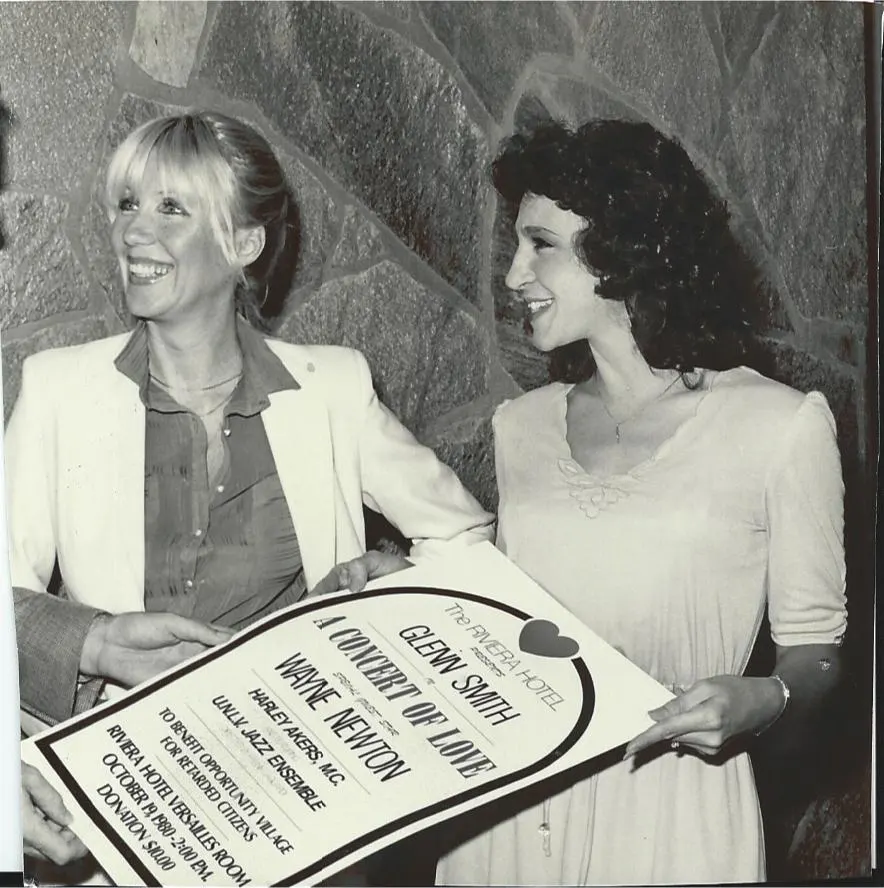 Two women smile while holding a sign for a charity event featuring Wayne Newton. The event supports Opportunity Village, highlighting Linda Smith's dedication to nonprofit fundraising and advocacy.| Linda’s Life – Linda Smith