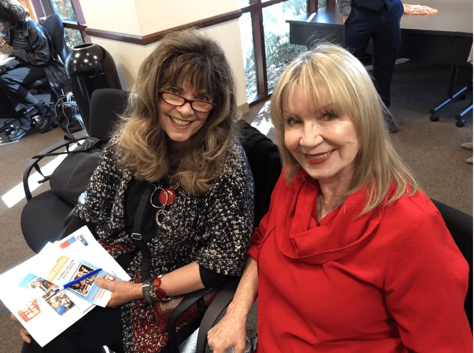 Two women sitting in a room, smiling warmly at the camera. One holds brochures related to nonprofit fundraising. The scene reflects advocacy and community engagement, highlighting the essence of motivational speaking and philanthropy.| Linda’s Life – Linda Smith