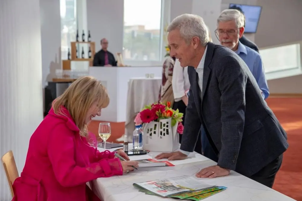 Linda Smith, a motivational speaker, signs a book for a man at a charity event. She is wearing a bright pink coat, sitting at a table with flowers and a wine glass. The setting is a warmly lit room with a few people in the background.| Linda’s Life – Linda Smith