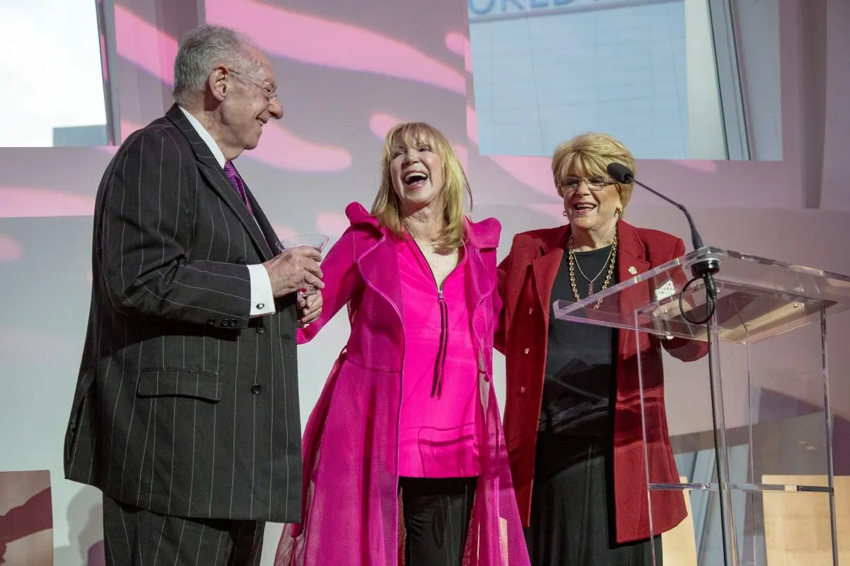 Linda Smith, a motivational speaker, stands smiling between two people at a charity event. The woman in pink is at a podium, engaging with the audience, highlighting her role in advocacy and nonprofit leadership.| Linda’s Life – Linda Smith