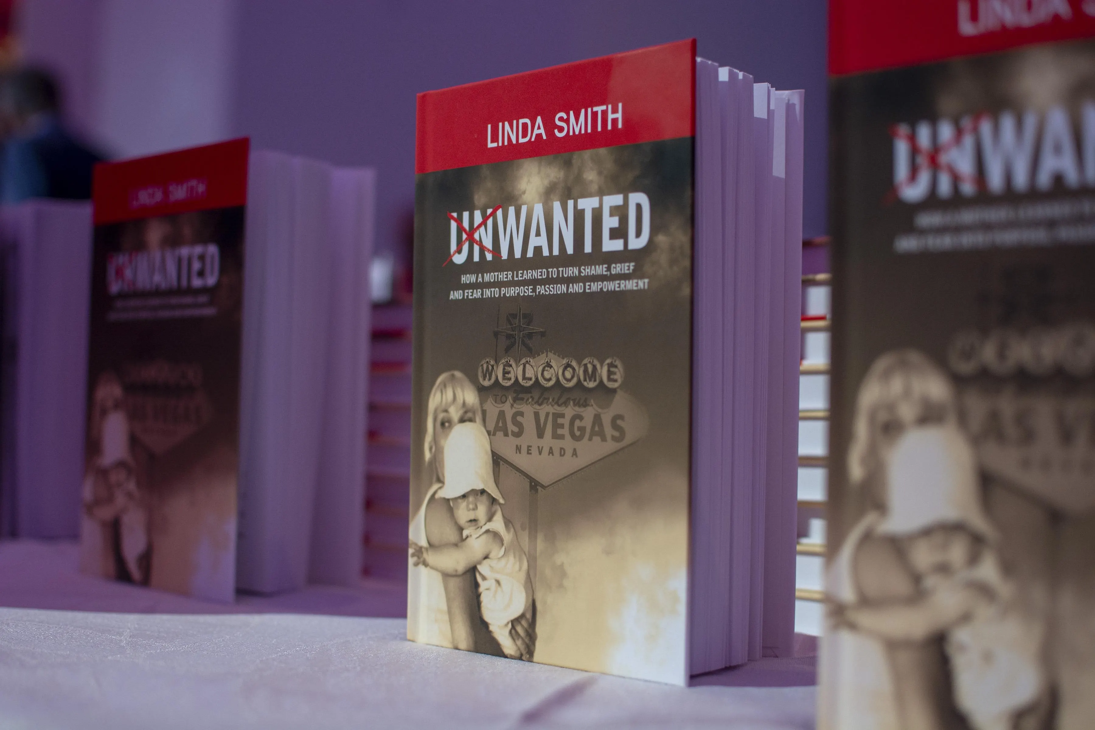 Several copies of "Unwanted" by Linda Smith are displayed upright on a table. The book cover shows a vintage photo of a woman holding a baby in front of a Las Vegas sign, highlighting her work in nonprofit fundraising and advocacy.| Linda’s Life – Linda Smith