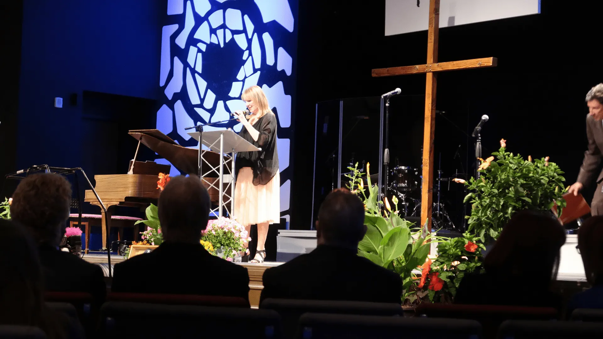 Linda Smith, a motivational speaker, stands at a podium on stage, addressing an audience in a hall decorated with plants and a piano, highlighting her advocacy and inspirational storytelling.| Linda’s Life – Linda Smith