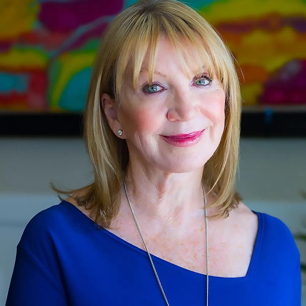 A woman in a vibrant blue top stands in front of a colorful abstract painting, smiling warmly. Her poised demeanor reflects her role as a motivational speaker and disability advocate, emphasizing her dedication to nonprofit leadership and advocacy.| Linda’s Life – Linda Smith