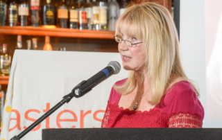 Linda Smith, a motivational speaker, addresses an audience at a charity event. She stands at a podium, speaking into a microphone, with a backdrop of shelves filled with bottles, emphasizing her advocacy and inspirational storytelling.| Linda’s Life – Linda Smith