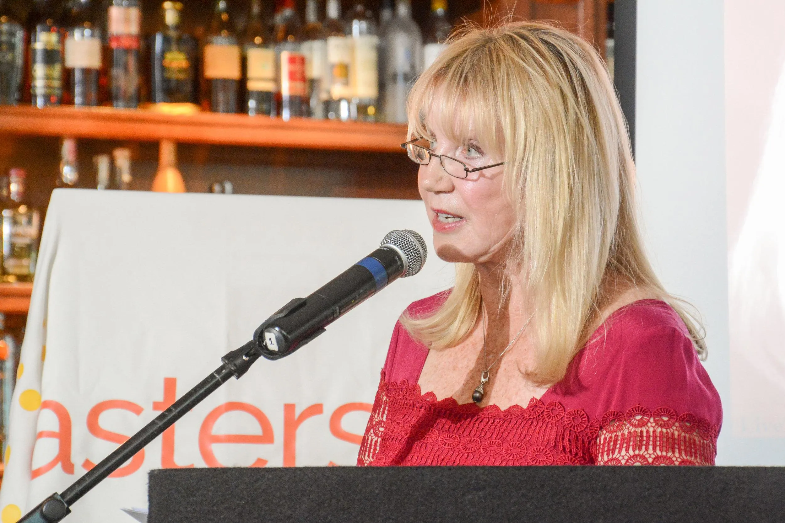 Linda Smith, a motivational speaker, addresses an audience at a charity event. She stands at a podium, speaking into a microphone, with a backdrop of shelves filled with bottles, emphasizing her advocacy and inspirational storytelling.| Linda’s Life – Linda Smith