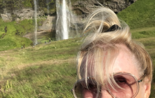 A woman smiles brightly in front of a lush green landscape with a waterfall, embodying the spirit of motivational speaker and disability advocate Linda Smith, known for her inspirational storytelling and advocacy in Down syndrome awareness.| Linda’s Life – Linda Smith