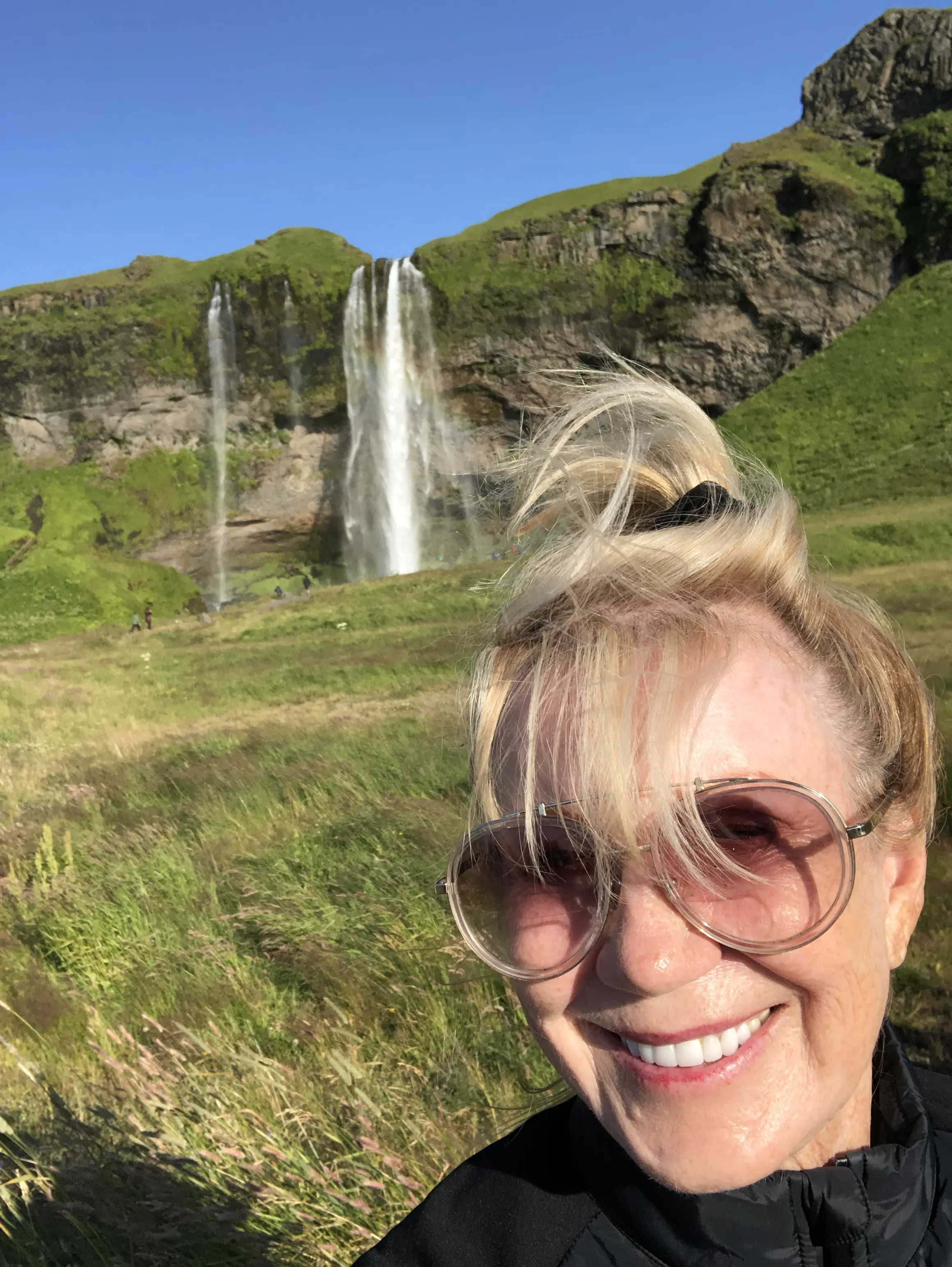 A woman smiles brightly in front of a lush green landscape with a waterfall, embodying the spirit of motivational speaker and disability advocate Linda Smith, known for her inspirational storytelling and advocacy in Down syndrome awareness.| Linda’s Life – Linda Smith