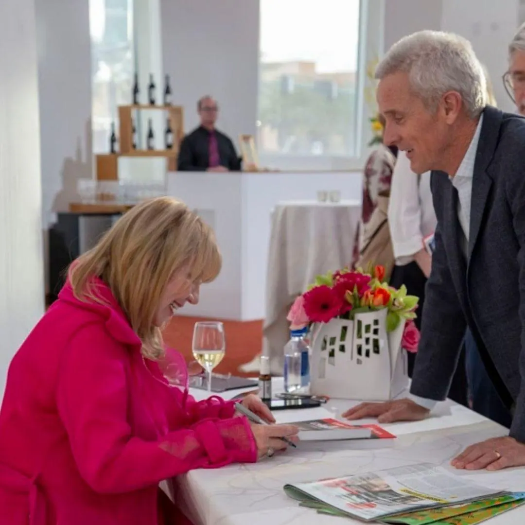 Linda Smith, a motivational speaker, signs a book at a charity event. She wears a bright pink jacket and smiles while interacting with a man. A table decorated with flowers and wine glasses adds to the lively atmosphere.| Linda’s Life – Linda Smith