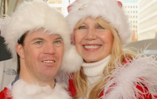 A woman and a man, both in festive Santa hats and red attire, smile warmly at a charity event focused on Down syndrome awareness, embodying the spirit of nonprofit fundraising and advocacy.| Linda’s Life – Linda Smith