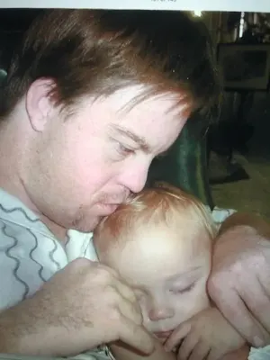 A man gently holds a sleeping baby in his arms, both resting comfortably. This tender moment reflects themes of love and care, resonating with Linda Smith’s advocacy for Down syndrome awareness and her commitment to disability advocacy.| Linda’s Life – Linda Smith