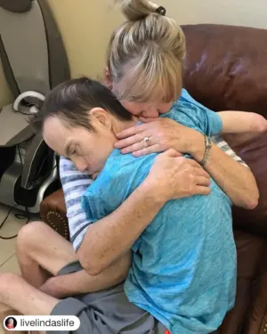 A woman tenderly hugs a man wearing a blue shirt, both seated on a brown couch. The moment captures a heartfelt connection, reflecting themes of Down syndrome awareness and motivational speaker Linda Smith's advocacy work in disability support.| Linda’s Life – Linda Smith