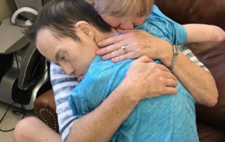 A woman tenderly hugs a man wearing a blue shirt, both seated on a brown couch. The moment captures a heartfelt connection, reflecting themes of Down syndrome awareness and motivational speaker Linda Smith's advocacy work in disability support.| Linda’s Life – Linda Smith