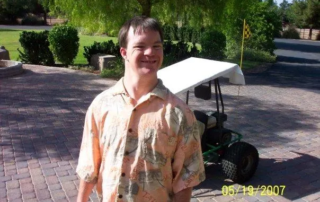 A young man with Down syndrome smiles brightly in a sunny driveway, wearing a patterned shirt. A golf cart is visible behind him. This image…| Linda’s Life – Linda Smith