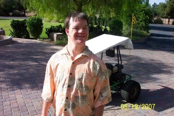 A young man with Down syndrome smiles brightly in a sunny driveway, wearing a patterned shirt. A golf cart is visible behind him. This image…| Linda’s Life – Linda Smith