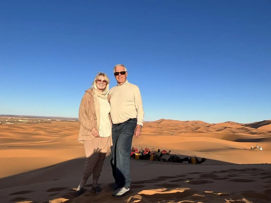 A couple stands smiling on sunlit sand dunes under a clear blue sky. Behind them, a group of people rests on the dunes. The scene evokes a sense of adventure and tranquility, reflecting themes of philanthropy and inspirational storytelling.| Linda’s Life – Linda Smith