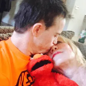 A young man wearing an orange shirt hugs a woman with blonde hair, both smiling gently. He holds a red Elmo plush toy. This tender moment reflects Linda Smith’s advocacy for Down syndrome awareness and inspirational storytelling.| Linda’s Life – Linda Smith