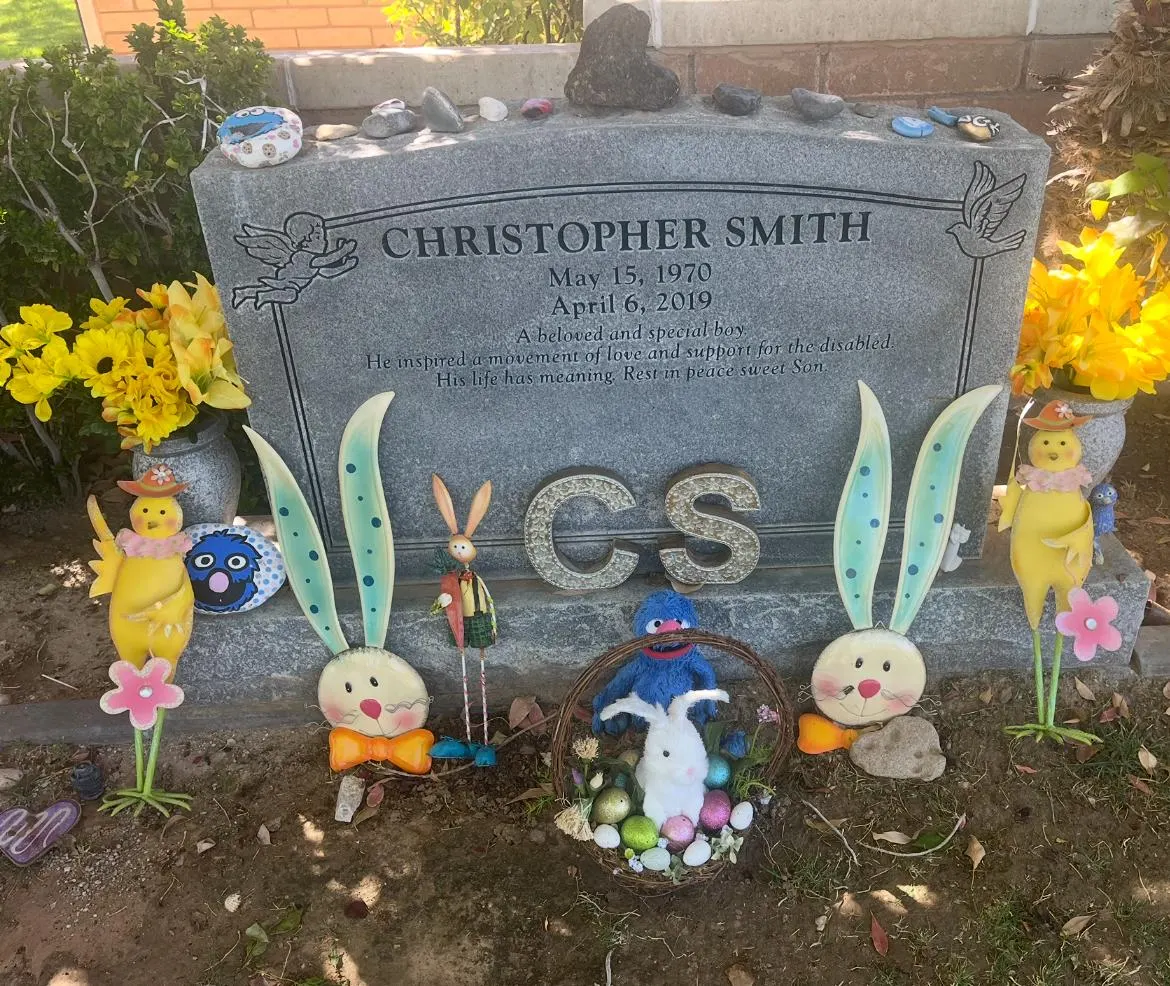 A gravestone for Christopher Smith is adorned with colorful decorations and flowers, including bunnies and chicks. This reflects Linda Smith's advocacy and nonprofit leadership in Down syndrome awareness and disability support.| Linda’s Life – Linda Smith
