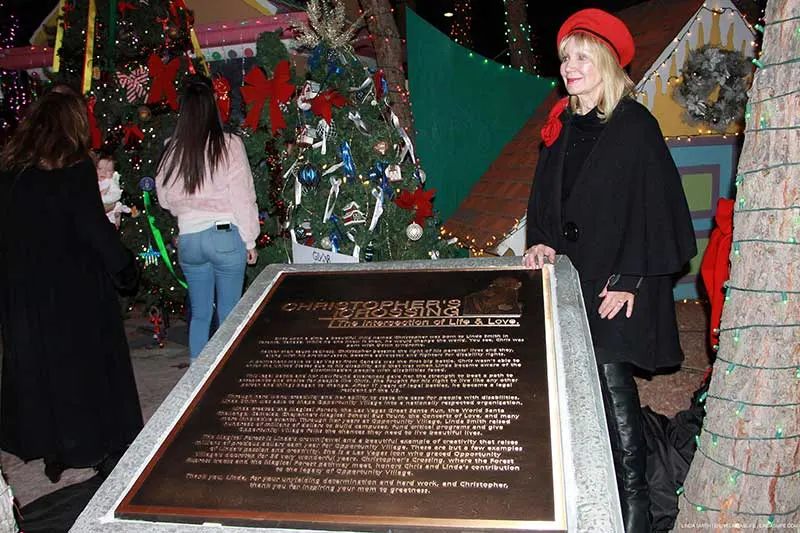 Linda Smith, a motivational speaker and disability advocate, stands beside a plaque at a charity event, surrounded by festive Christmas trees adorned with ornaments and lights, highlighting her commitment to philanthropy and nonprofit fundraising.| Linda’s Life – Linda Smith