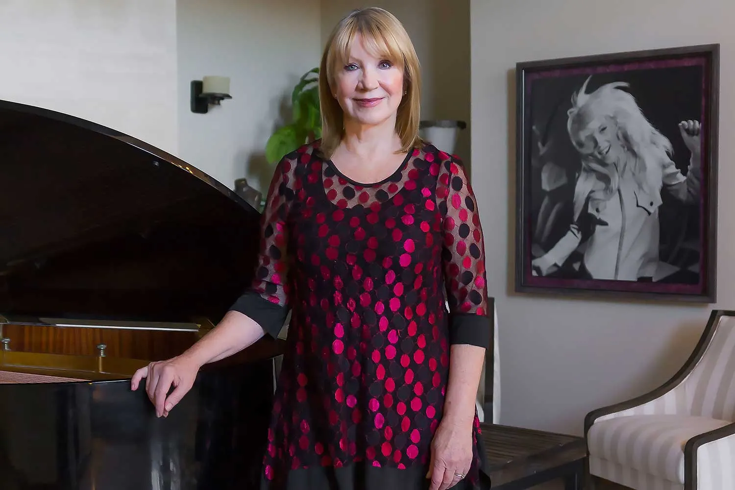 A woman in a red and black polka dot top stands beside a grand piano, with a framed photo behind her. The setting is elegant, reflecting themes of philanthropy and nonprofit leadership.| Linda’s Life – Linda Smith