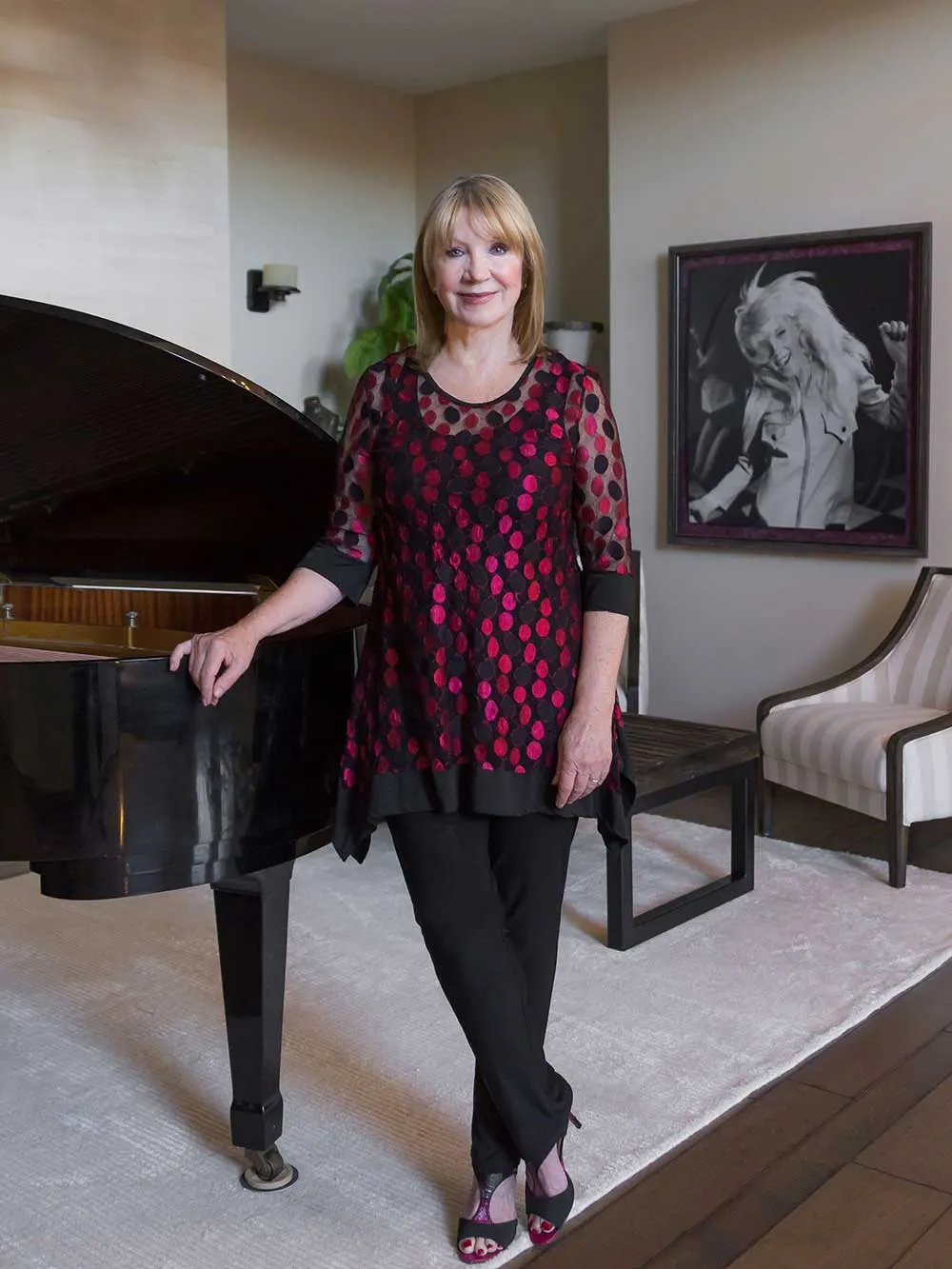 A woman in a red and black polka dot top stands beside a grand piano in a modern room. A black-and-white portrait hangs on the wall. This image represents nonprofit leadership and advocacy, highlighting her role as a disability advocate.| Linda’s Life – Linda Smith